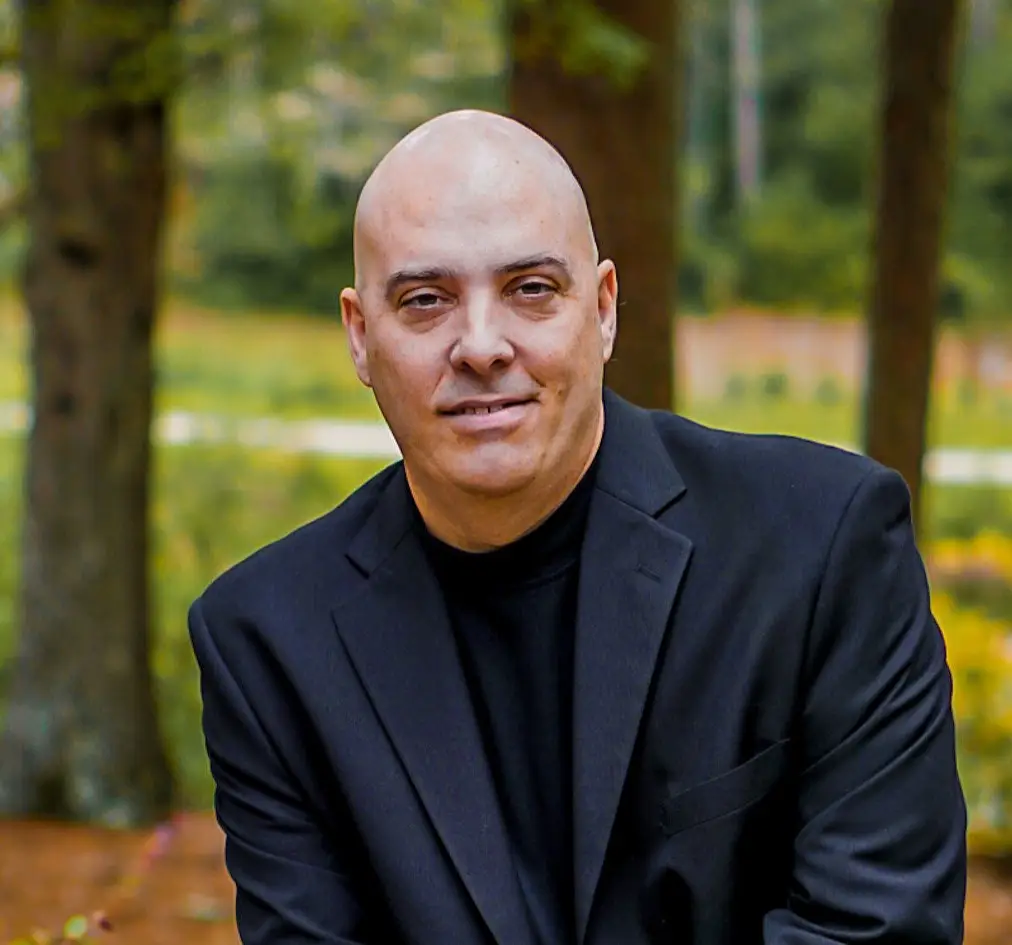 About Us 3 a man in a black suit standing in a park with tall trees and green grass in the far background