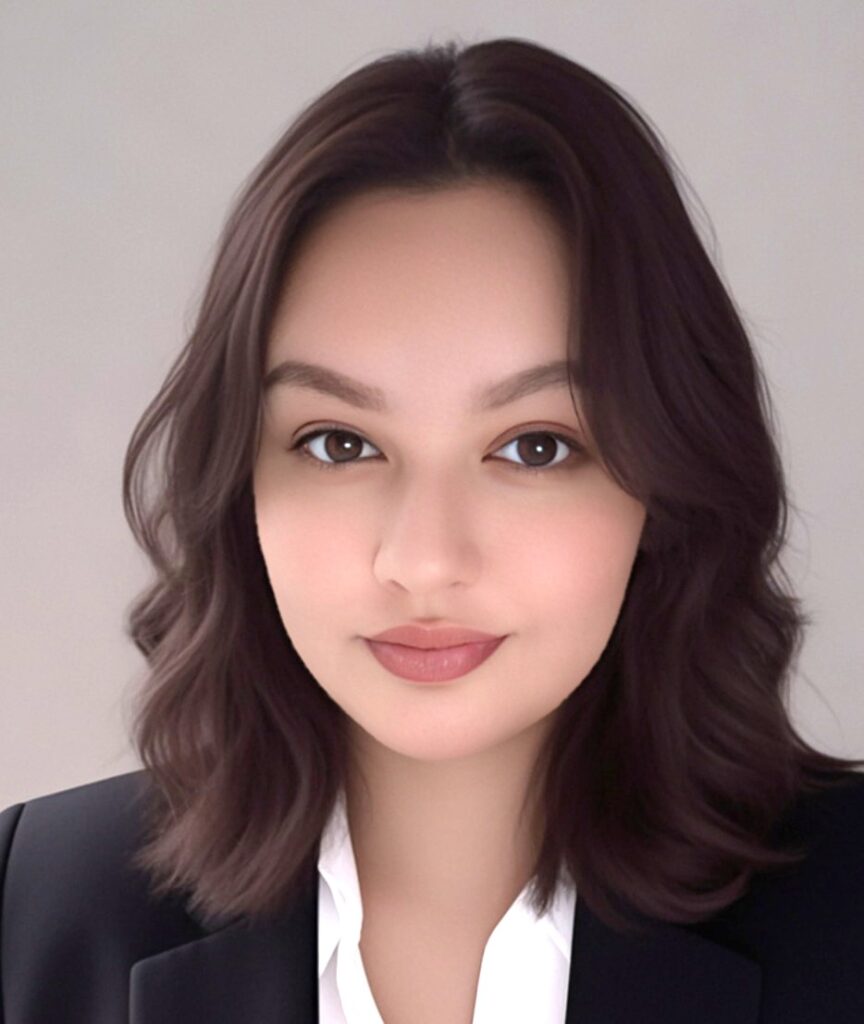About Us 9 Portrait of a person with shoulder-length wavy dark hair, wearing a black blazer and white shirt, against a neutral background.